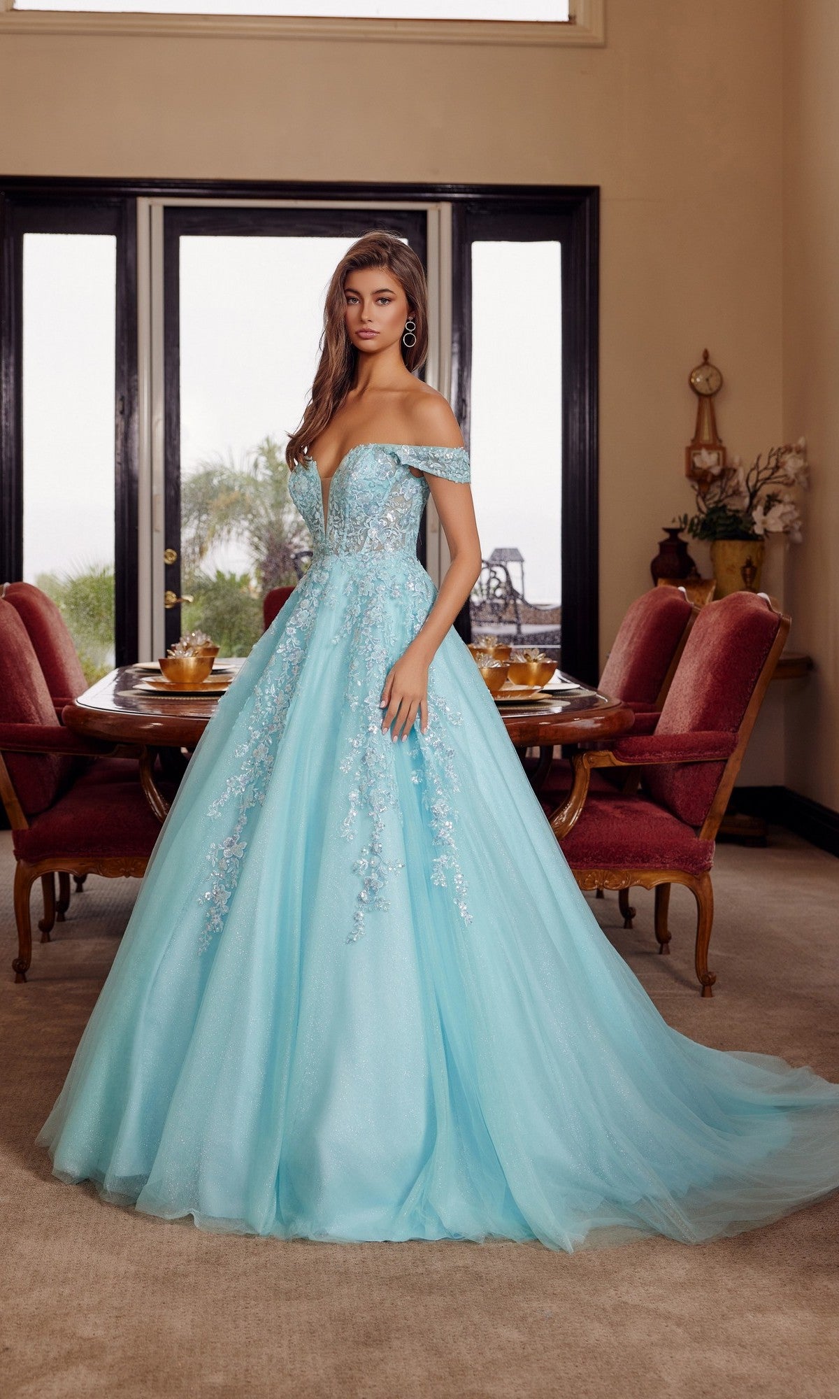 A woman wearing the aqua Long Prom Dress: Nox Anabel H1881, an off-the-shoulder tulle ball gown with floral embellishments, stands in an elegant dining room with red chairs and a round wooden table.